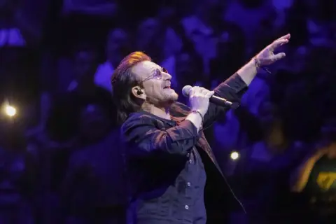 Bono z U2 śpiewa na koncercie, zapowiadając u2 nowa płyta. W tłumie widać światła i sylwetki fanów.