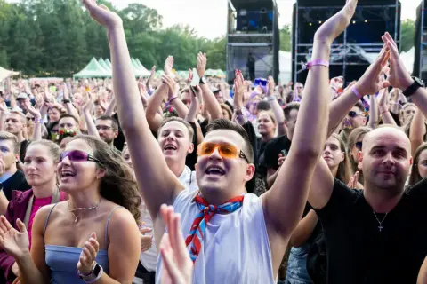 Tłum na koncercie cieszy się letnie brzmienia. Ludzie klaszczą, podnoszą ręce, a niektórzy mają kolorowe okulary i chusty.