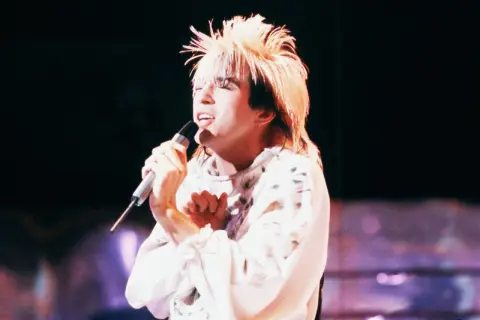 Limahl piosenkarz z charakterystyczną fryzurą śpiewa do mikrofonu na scenie.