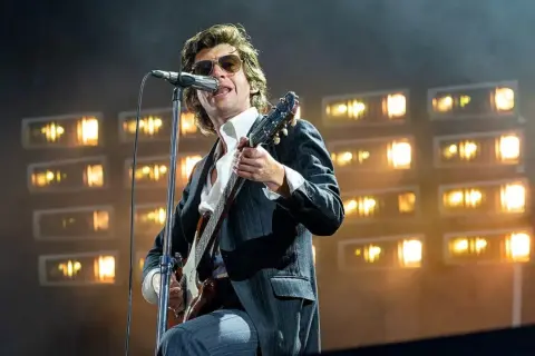 Alex Turner z Arctic Monkeys na scenie, w okularach i garniturze, gra na gitarze podczas jednego z niezapomnianych koncertów.