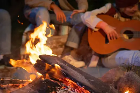 Przy ognisku siedzą ludzie, grają na gitarze i śpiewają piosenki na ognisko.