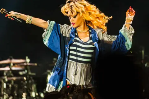 Rihanna na koncercie w Polsce, w bluzie z kolorowymi pasami, śpiewa z mikrofonem w dłoni.