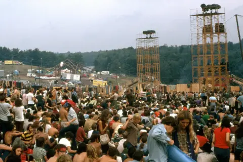 Tłum ludzi na festiwalu Woodstock, z widocznymi wieżami i sceną w tle.