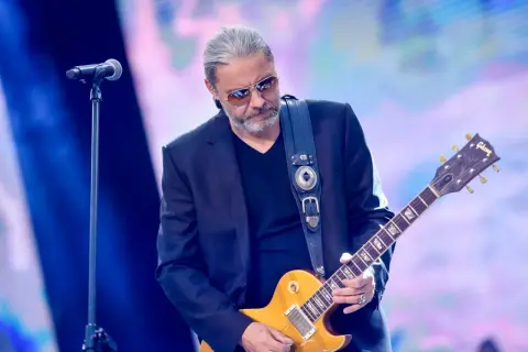 Dżem wokalista z gitarą Gibson na scenie, w okularach przeciwsłonecznych, z siwymi włosami.