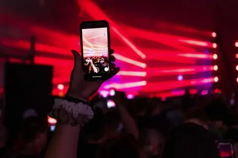 Dłoń trzyma smartfon nagrywający koncert z czerwonymi światłami w tle.