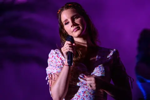 Lana Del Rey śpiewa na scenie w kwiecistej sukience, oświetlona fioletowym światłem.