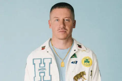 Macklemore w białej kurtce z naszywkami, gotowy na macklemore koncert polska.