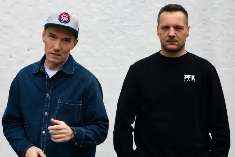 Dwóch członków Paktofoniki, jeden w czapce z daszkiem i granatowej kurtce, drugi w czarnej bluzie z logo PFK.