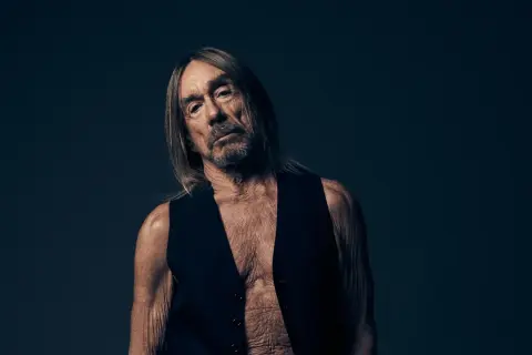 Iggy Pop w czarnej kamizelce, z długimi włosami i zmęczoną miną, na ciemnym tle.