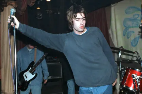 Liam Gallagher z Oasis śpiewa na scenie, w tle basista i perkusja. To klasyczne oasis utwory.
