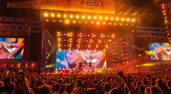 Tłum na festiwalu, scena z wielkim ekranem i logo "FEST festival program". Światła i artyści na scenie.