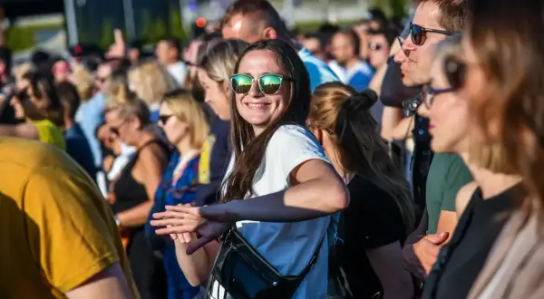 Uśmiechnięta kobieta w okularach przeciwsłonecznych na tle tłumu ludzi na koncercie.