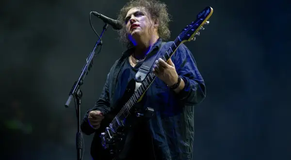 Robert Smith z The Cure na koncercie, w charakterystycznym makijażu, gra na czarnej gitarze elektrycznej.