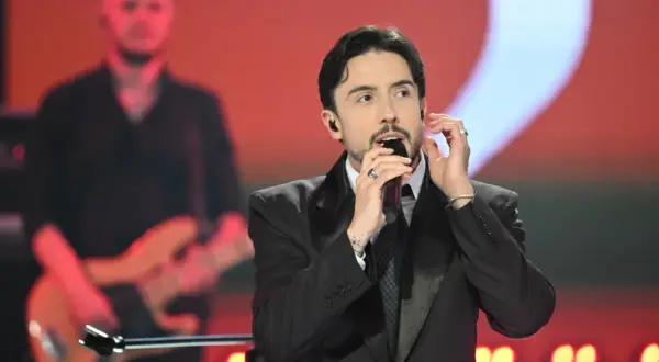 Dawid Kwiatkowski w czarnym garniturze śpiewa do mikrofonu na scenie. W tle muzyk z gitarą.