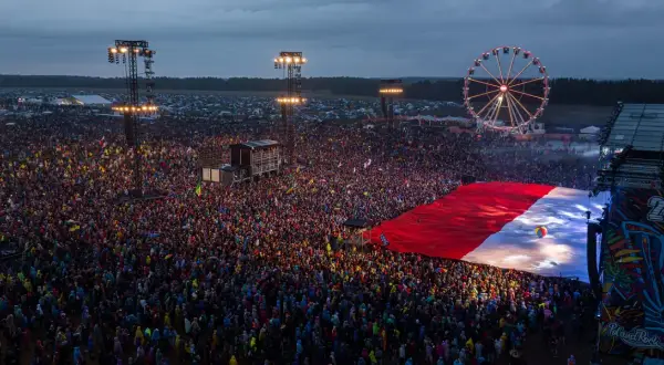 Ogromny tłum ludzi na festiwalu rockowym, rozłożona flaga Polski, scena i diabelski młyn.