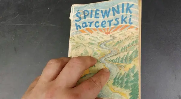 Dłoń trzyma stary śpiewnik harcerski z rysunkiem rzeki i gór.