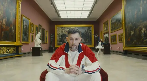 Mężczyzna w dresie siedzi w galerii sztuki, otoczony obrazami i rzeźbami.