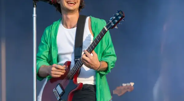 Joe Keery w zielonej koszuli gra na czerwonej gitarze elektrycznej na scenie.