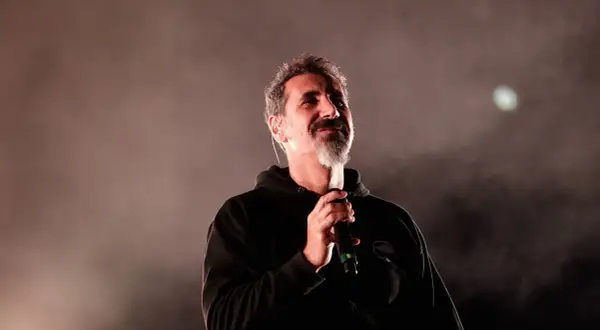 Serj Tankian z System of a Down na koncercie, trzyma mikrofon, w tle mgła i światło.