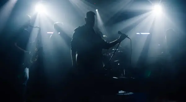 Zespół muzyczny na scenie, wokalista z mikrofonem, saksofonista, światła sceniczne.