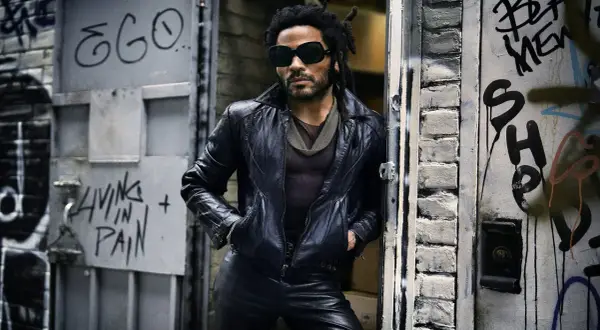 Lenny Kravitz w skórzanej kurtce i okularach, oparty o drzwi z graffiti, jakby właśnie wysiadł z łodzi.