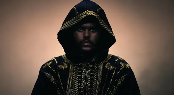 The Weeknd w czarnej, haftowanej złotem kurtce z kapturem, patrzący prosto w obiektyw.
