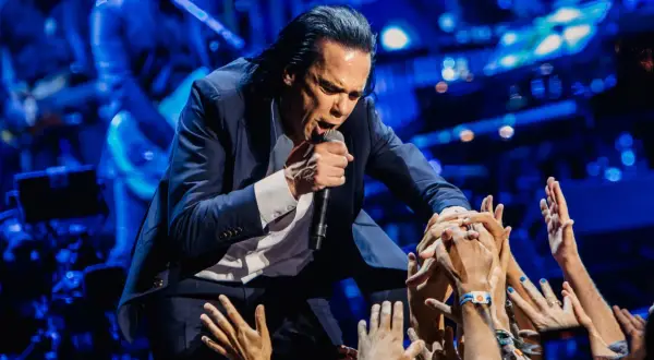 Nick Cave w garniturze śpiewa do mikrofonu, otoczony przez wyciągnięte ręce fanów.