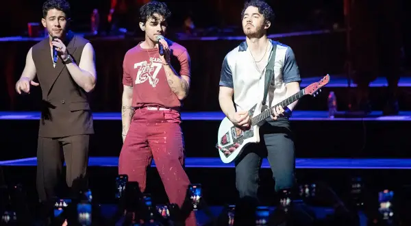 Trzech członków zespołu Jonas Brothers na scenie, jeden śpiewa, drugi gra na gitarze.