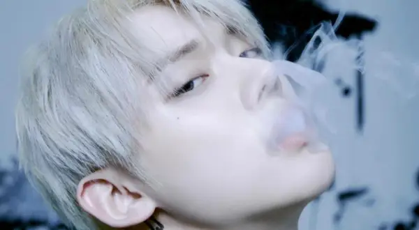 Młody mężczyzna z jasnymi włosami, jeden z członków popularnych k-pop zespołów męskich, wydmuchuje dym.