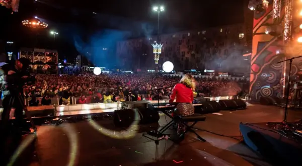 Koncert na scenie z klawiszowcem, tłum ludzi na placu, kamery, światła i dym.