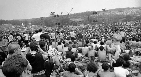 Tysiące ludzi siedzi na trawie, patrząc na scenę festiwalu. To właśnie Woodstock, gdzie jest to legendarne wydarzenie.