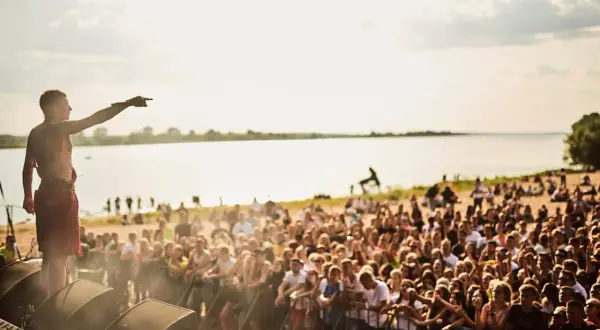 Artysta na scenie podczas jednego z letnich festiwali w Polsce, wskazujący na tłum zgromadzonych fanów nad jeziorem.