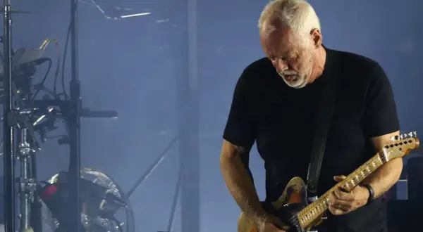 David Gilmour gra na gitarze elektrycznej na tle sceny z dymem i sprzętem muzycznym.