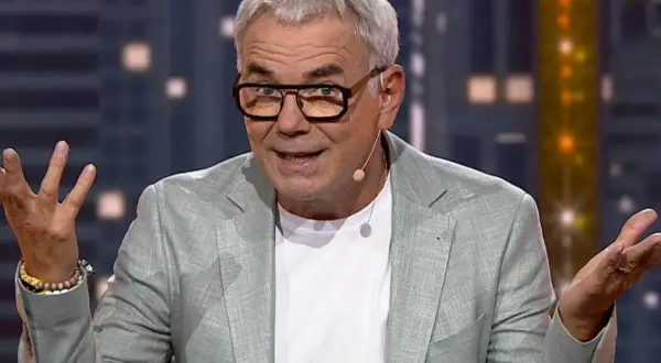 Mężczyzna w okularach i jasnej marynarce gestykuluje, jakby śpiewał z playbacku.