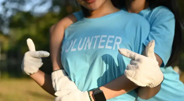 Dwie młode osoby w niebieskich koszulkach z napisem "VOLUNTEER" i białych rękawiczkach, pozujące na tle zieleni.