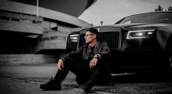 Mężczyzna w czapce i kurtce siedzi obok czarnego Rolls-Royce'a na tle betonowej budowli.