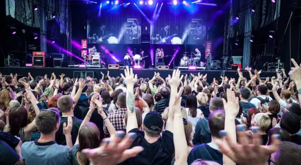 Koncert na żywo, tłum ludzi z podniesionymi rękami, scena z zespołem w tle, fioletowe światła.