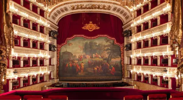 Wnętrze luksusowego teatru operowego z czerwonymi lożami, złotymi zdobieniami i malowidłem na scenie.
