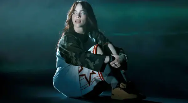 Billie Eilish siedzi na scenie w stroju sportowym, wpatrując się w bok.
