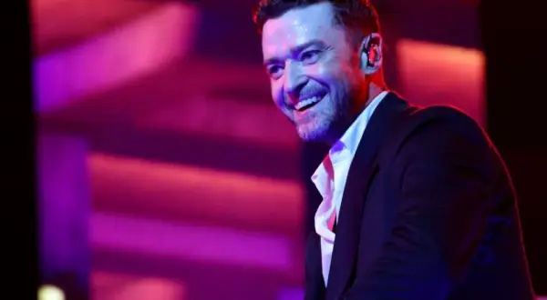 Justin Timberlake w czarnym garniturze i białej koszuli, uśmiecha się na tle fioletowo-różowego oświetlenia scenicznego.
