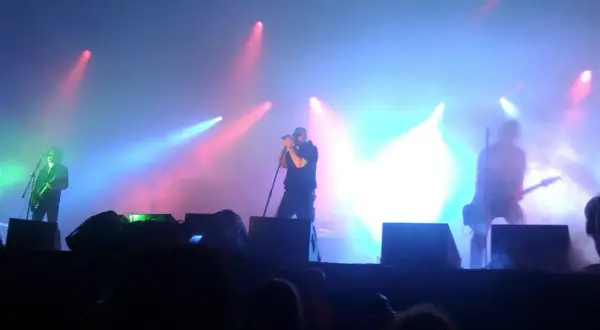 Na scenie podczas koncertu Sisters of Mercy, w dymie i kolorowych światłach, artyści grają na gitarach i śpiewają.