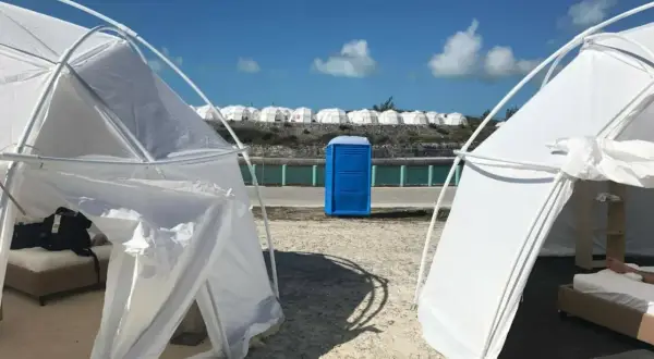 Namioty i niebieska toaleta polowa na plaży, przypominające fiasko Fyre Festival 2. W tle rzędy białych kopuł.