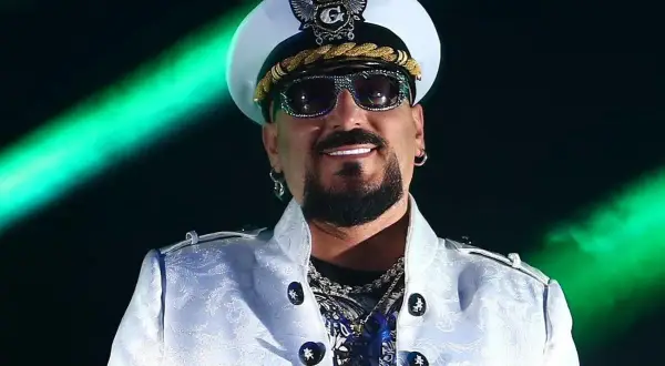Gigi D'Agostino w białej marynarce i czapce, w okularach przeciwsłonecznych, na tle zielonych świateł.