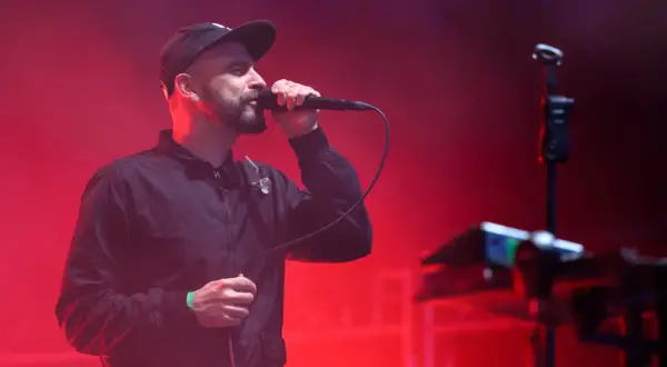 Wokalista z brodą w czapce i kurtce śpiewa do mikrofonu na tle czerwonych świateł. To część tauron nowa muzyka line-up.