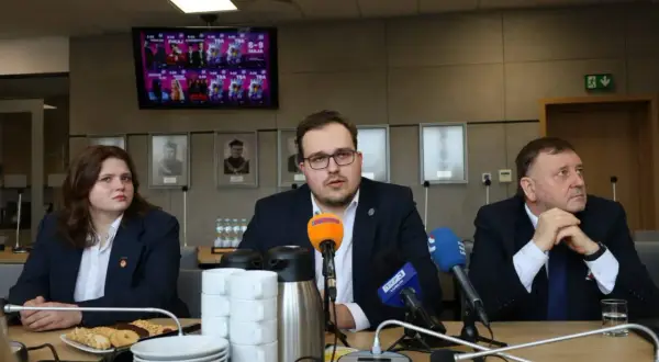 Trzy osoby siedzą przy stole konferencyjnym z mikrofonami i kubkami. Na ekranie w tle widnieje plakat.