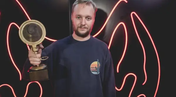 Mężczyzna w granatowej bluzie trzyma złotą statuetkę na tle czerwonych neonów.