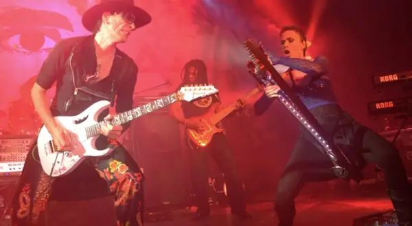 Trzech gitarzystów na scenie, jeden w kapeluszu gra na białej gitarze, drugi w tle na pomarańczowej, trzeci na futurystycznej.