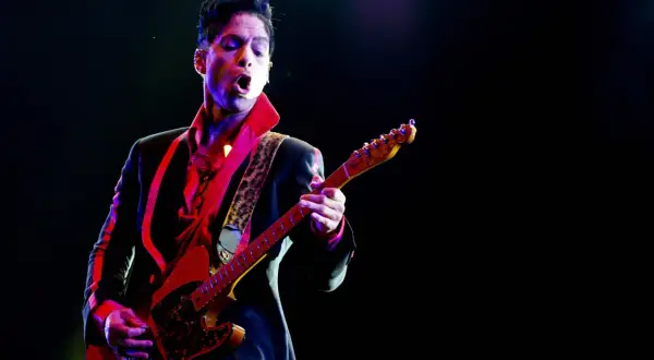 Prince, jako prince opener, gra na gitarze w czerwonej koszuli i czarnej marynarce, oświetlony kolorowymi światłami sceny.