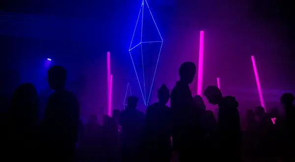 Sylwetki ludzi na tle niebiesko-fioletowych świateł i geometrycznej instalacji świetlnej.