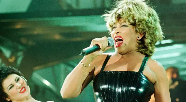 Tina Turner śpiewa do mikrofonu na scenie, w błyszczącej czarnej sukience.